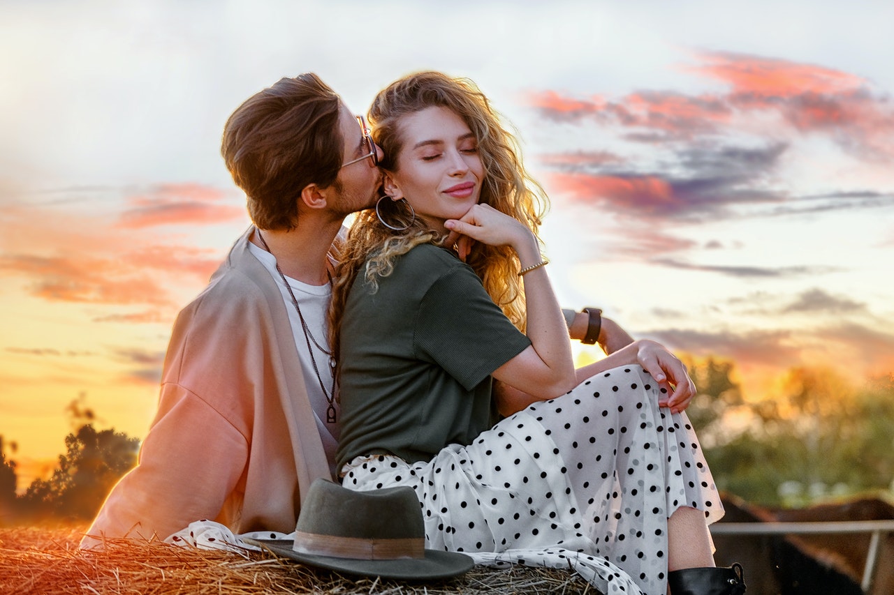 Man kissing his attractive girlfriend while sitting on a haystack during sunset